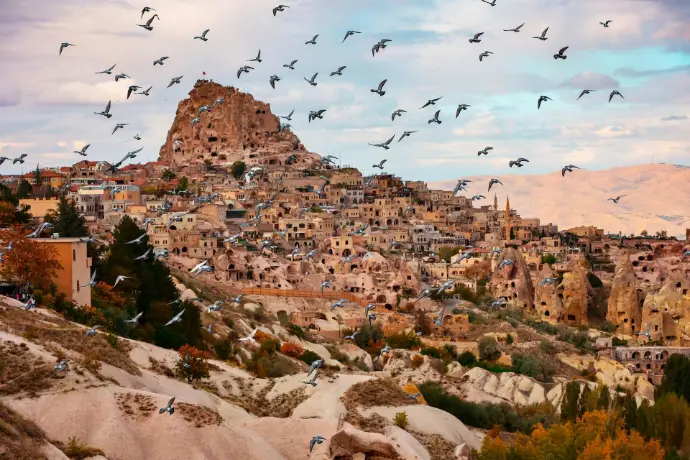 Pigeons flying over Pigeon Valley with Uchisar Castle and stone houses in Cappadocia, Turkey.